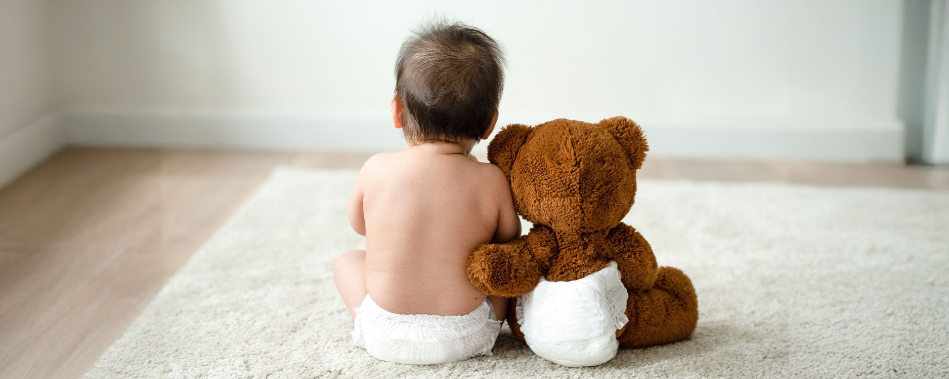 Bebé sentado en el suelo junto a un peluche, ambos con pañal, representando el cuidado de la dermatitis del pañal